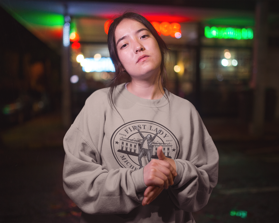 A young woman wearing a light gray sweatshirt with a black graphic print that reads 'FIRST LADY MICHELLE OBAMA' stands outdoors at night in front of a blurred storefront with colorful neon lights.